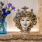 Medusa head with flowers on a stone shelf beside a vase of blue irises