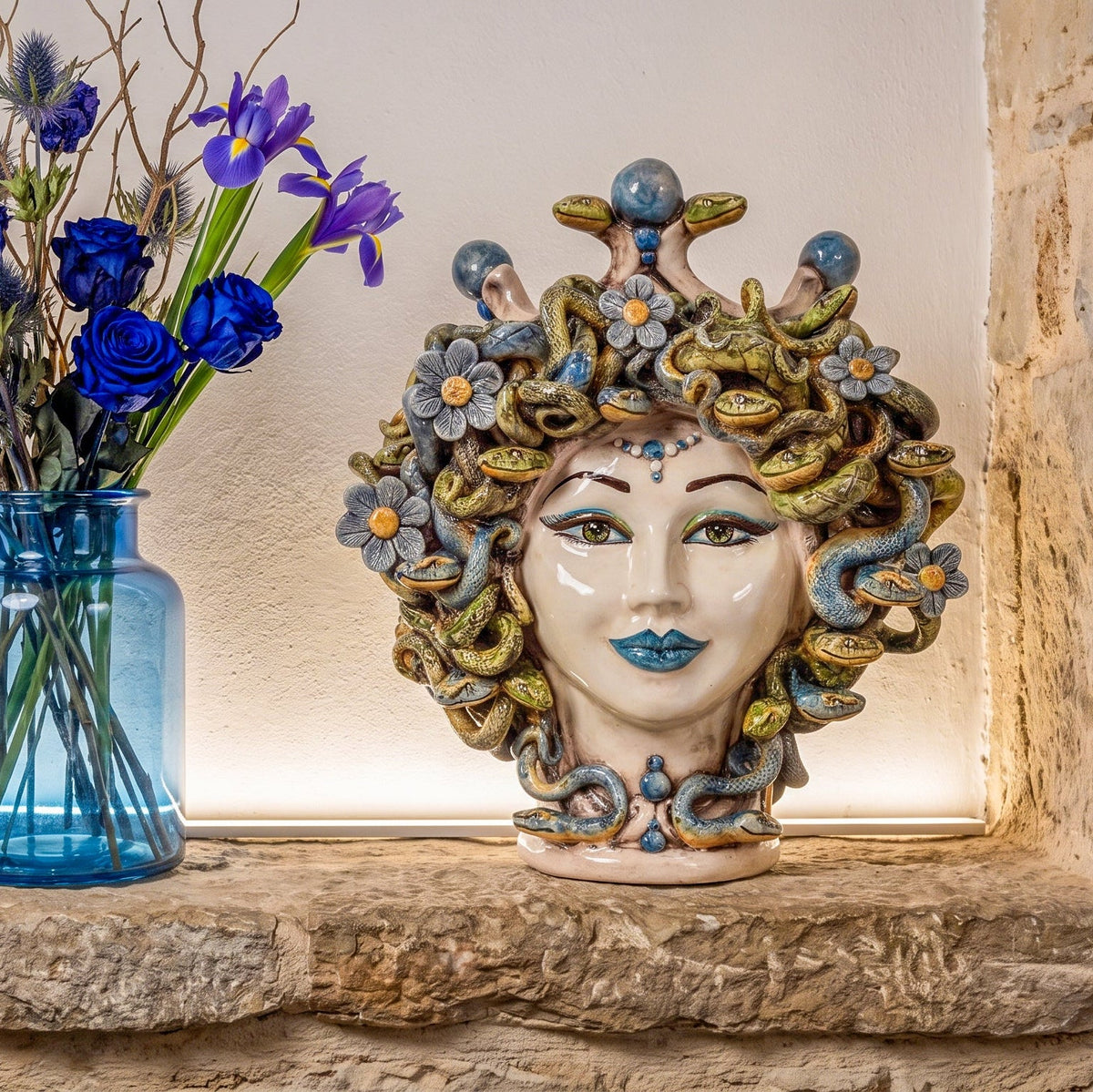 Medusa head with flowers on a stone shelf beside a vase of blue irises