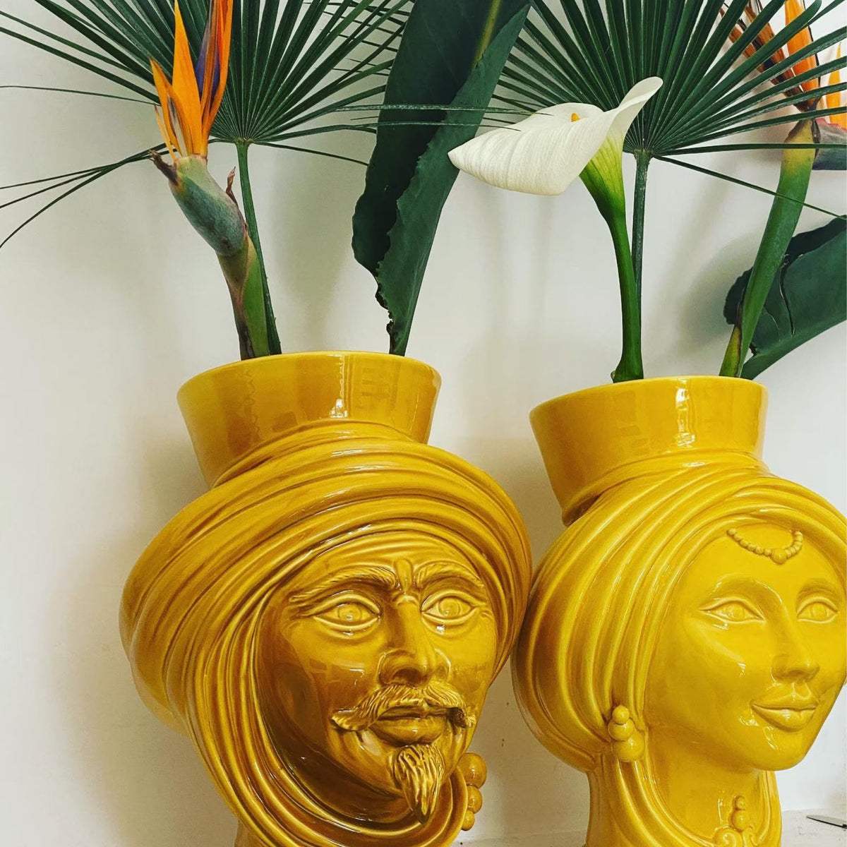 Pair of mustard Teste di Moro, bearded man and woman, used as vases with bird-of-paradise flowers and calla lilies